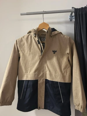 Beige och marinblå vindjacka från Hummel - Snygg vindjacka från Hummel i beige och marinblått med huva och dragkedja. Jackan har vattenavvisande och ventilerande material, samt praktiska fickor med vita detaljer. Perfekt för dig som vill ha en stilren och funktionell jacka till vardags. Mitt namn står skrivet på jackan.