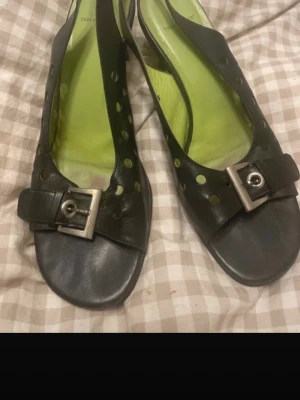 Svarta sandaler från Ten Points - Säljer ett par svarta sandaler från Ten Points i skinn med coola håldetaljer och en stor metallspänne framtill. Insidan är limegrön för en extra pop av färg. Öppen tå och låg klack gör dem perfekta för varma dagar.