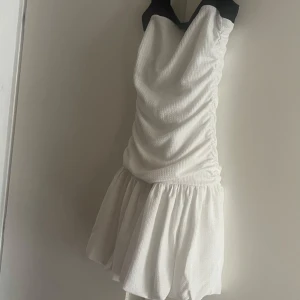 Vit klänning med svart inslag från Gina Tricot - Supersöt vit kort klänning från Gina Tricot med svart kant upptill. Klänningen har en rynkad överdel och volangig kjol, perfekt för dig som gillar en feminin och trendig look. Materialet är strukturerat och känns lätt mot huden. Perfekt på en fest med en bolero eller kavaj eller att använda utomlands i värmen.