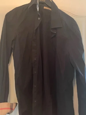 Svart skjorta från Burberry  - Snygg svart skjorta från Burberry i storlek L. Den har långa ärmar och klassiska rutiga detaljer på insidan av kragen och ärmsluten. Perfekt för både vardag och fest! Skjortan är bra skick och har knappar framtill. Hör av dig vid vid minsta fråga eller fundering! 😃 