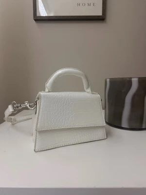 Vit croco handväska med axelrem - Snygg liten handväska i vit färg med croco-mönster och silverfärgade detaljer. Väskan har både handtag och avtagbar axelrem, samt två fack inuti. Perfekt för att bära det viktigaste och ge din outfit en clean vibe.