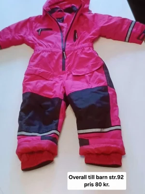 Rosa täckbyxor med svarta detaljer - Säljer ett par rosa täckbyxor med svarta förstärkningar på knän och ben, samt reflexdetaljer. Byxorna har dragkedja framtill och är gjorda i slitstarkt syntetmaterial, perfekta för kalla dagar utomhus.