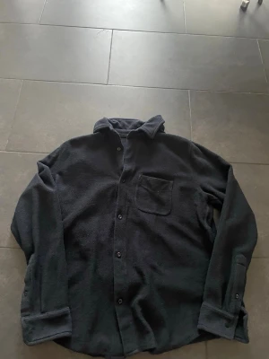 Svart overshirt med knappstängning - Svart overshirt med mjukt fleece-material och klassisk krage. Jackan har knappstängning framtill och en bröstficka på vänster sida. Perfekt att slänga på över en hoodie eller t-shirt för en chill look.