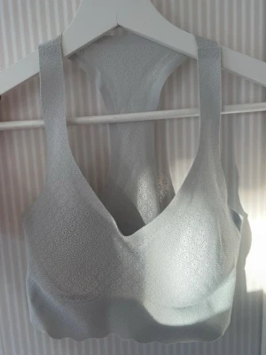 Ljusgrå brottarrygg-bh från Sloggi - Superskön ljusgrå bh från Sloggi med brottarrygg och diskret hålmönster över hela tyget. Västen är sömlös och har breda axelband för extra komfort. Perfekt för dig som gillar en enkel men snygg look.