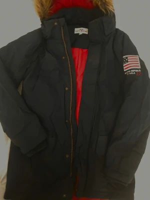 Mörkblå parkas från Hampton Republic - Säljer en mörkblå parkas från Hampton Republic med röd innerfoder och avtagbar huva med fuskpäls. Jackan har dragkedja och knappar framtill, samt amerikansk flagga som patch på ärmen. Perfekt för kalla dagar och riktigt snygg streetstil.