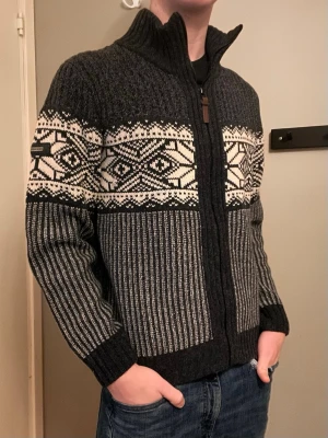 Mörkgrå stickad cardigan med mönster - Säljer en mörkgrå stickad kofta från märket Pierre cardigan Paris, med dragkedja och hög krage. Koftan har ett snyggt vitt och svart nordiskt mönster över bröstet och ribbade partier på ärmar och nedre delen. Perfekt för kalla dagar och riktigt stilren. Allt gott deluxe garderoben 🤩