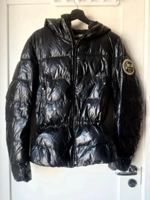 Svart dunjacka från Kejo  - Säljer en svart glansig dunjacka från Kejo med huva och dragkedja. Jackan har broderad Kejo-logga på ärmen och stor brodyr på ryggen i guld och vitt. Puffig modell med markerad midja och långa ärmar. Fylld med äkta gåsdun för extra värme.
