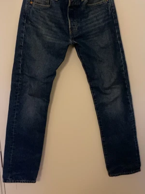 Levi's 501 jeans blå denim 29/32 - Klassiska Levi's 501 jeans i mörkblå denim med raka ben och fem fickor. Jeansen har en brun läderpatch bak i midjan och den ikoniska röda Levi's-taggen på bakfickan. Materialet är slitstarkt bomullsjeans och modellen har normal passform.