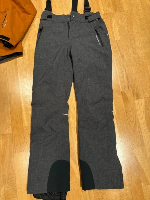 Mörkgrå täckbyxor med hängslen - Säljer ett par mörkgrå täckbyxor från Didriksons med justerbara svarta hängslen och förstärkta partier vid bensluten. Byxorna har dragkedjor vid fickorna och reflexdetaljer på benen. Perfekta för vintern och outdoor-aktiviteter.