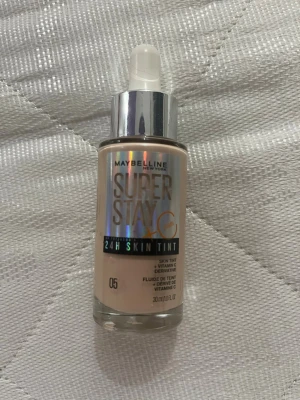 Maybelline Super Stay 24H Skin Tint 05 - Flytande foundation från Maybelline i nyansen 05, Super Stay 24H Skin Tint med vitamin C. Kommer i en genomskinlig glasflaska med silvrig pump och holografisk etikett. Perfekt för en naturlig och fräsch look med lätt täckning.