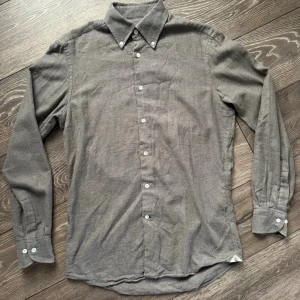 Grå skjorta från Morris Heritage - Snygg grå skjorta från Morris Heritage i mjuk bomull med diskret fiskbensmönster. Klassisk krage med knappar, långa ärmar och vita knappar framtill. Broderad detalj vid manschetten och liten logga vid nederkanten. Perfekt till jeans eller chinos. Den är rätt nopprig, därav priset