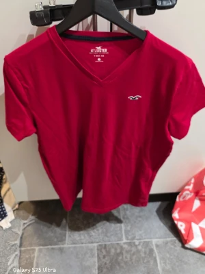 Röd v-ringad t-shirt från Hollister - Säljer en röd t-shirt från Hollister med v-ringad hals och korta ärmar. T-shirten har en liten broderad logga på bröstet och är gjord i mjuk bomull. Perfekt för dig som gillar enkel och stilren design med en touch av street.
