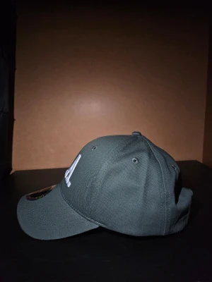 New Era - Snygg grön keps från Statewear med broderad vit L.A. text framtill. Klassisk böjd skärm och justerbar rem baktill för perfekt passform. Kepsen är tillverkad i slitstark bomull och har ventilerande hål upptill. Perfekt accessoar för en chill streetwear-look. Klistermäket och ramen är fortfarande på och kepsen är helt oanvänd!