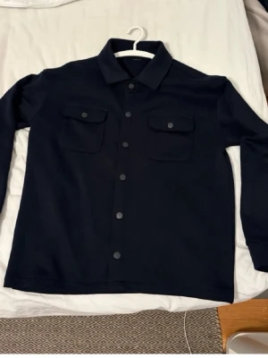 Mörkblå overshirt med knappar - Snygg mörkblå overshirt med klassisk krage och tryckknappar framtill. Jackan har två stora bröstfickor med lock och långa ärmar. Perfekt att slänga på över en hoodie för en chill look. Materialet känns. Strl S