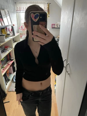 Svart croppad ribbad polotopp - Säljer en svart croppad polotopp med lång ärm och krage. Toppen är ribbad och har en djup v-ringning framtill. Snygg passform som sitter tight och visar midjan. Perfekt att styla med jeans eller kjol för en trendig look.