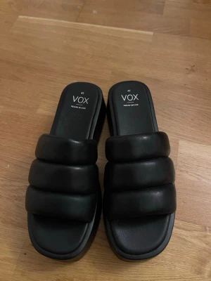 Svarta plattformssandaler från VOX - Chunky svarta sandaler från VOX med breda, vadderade remmar och fyrkantig tå. Plattformssula ger extra höjd och cool vibe. Tillverkade i veganskt material och har en stilren look som passar till många outfits.