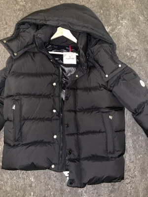 Svart Moncler pufferjacka med huva - Säljer en svart pufferjacka från Moncler med stor huva och klassisk Moncler-logga på ärmen. Jackan har dragkedja och knappar framtill, två sidofickor och quiltad design. Tillverkad i polyester och dun, perfekt för kalla dagar. Snygg och tidlös modell.