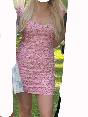 Blommig rosa kortklänning från Nelly -  kortklänning från Nelly i rosa med blommigt mönster. Klänningen har puffiga långärmar, rynkade detaljer och snörning i ryggen. Storlek 36, använt 1 gång. Säljer då den inte kommer till användning längre!