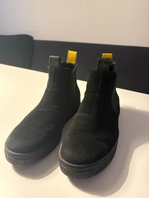  Canada Snow boots  - Svarta Chelsea boots från Canada Snow med grov platt sula och rund tå. Ovandel i mocka och detaljer i skinn, samt gul dragflik bak för extra stilpoäng. Perfekt för dig som gillar klassiska boots med en twist.
