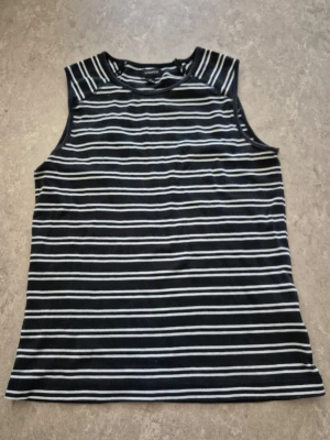 Linnetopp - Säljer en svart och vit randig linnetopp från Lindex i storlek S. Toppen är ärmlös med rund hals och har ett klassiskt, smalt randigt mönster. Perfekt för varma dagar och enkel att matcha med jeans eller shorts.