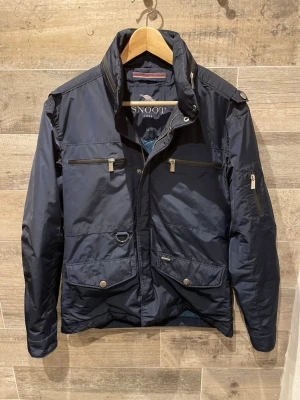 Snoot jacka  - Snygg mörkblå vindjacka från Snoot med flera dragkedjeförsedda fickor, hög krage och coola detaljer som axelklaffar och metallknappar. Jackan är tillverkad i polyester och har en modern, normal passform. Perfekt för dig som vill ha en stilren och funktionell jacka.