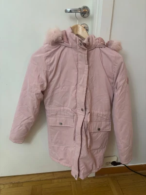 Ljusrosa parkas från Threadgirls - Säljer en ljusrosa parkas från Threadgirls med huva och fluffig fuskpälsdetalj. Jackan har dragsko i midjan, stora fickor med knappar och stängs med dragkedja och tryckknappar. Perfekt för kalla dagar och riktigt mysig att bära.
