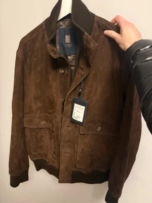 Mörkbrun mocka bomberjacka 1797 - Säljer en mörkbrun bomberjacka i mocka från 1797. Jackan har ribbade muddar, knappar framtill och två stora fickor med lock. Snygg och stilren modell som ger en lyxig känsla. Perfekt för dig som vill ha en klassisk jacka med modern touch.