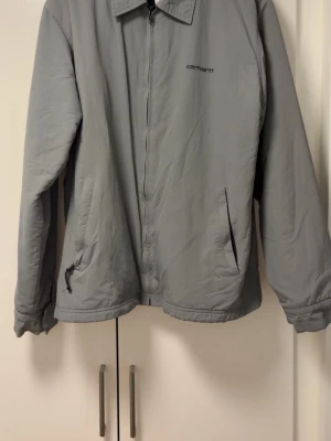 Grå vindjacka från Carhartt - Carhartt jacka vet inte storlek tyvärr passar L/M är i bra skick har själv thriftat den för 3 år sen och använt den ksk 2 gånger sen dess därav jag säljer