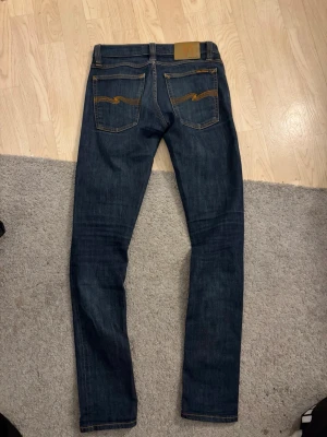 Mörkblå Nudie Jeans - Säljer ett par mörkblå jeans från Nudie Jeans med klassiska orange sömmar och broderade bakfickor. Jeansen har normal passform i midjan och är tillverkade i slitstarkt jeansmaterial av bomull. Perfekta för dig som gillar en smal siluett och tidlös stil.