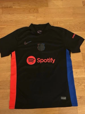 Svarta Nike fotbollströja Barcelona - Barcelona fotbollströja från Nike i svart med röda och blå detaljer på sidorna. Tryck med Spotify-logga på framsidan och Lamine Yamal 19 på ryggen. Tröjan har klubbmärke och UNHCR-logga, samt korta ärmar och krage. Perfekt för dig som älskar Barca.
