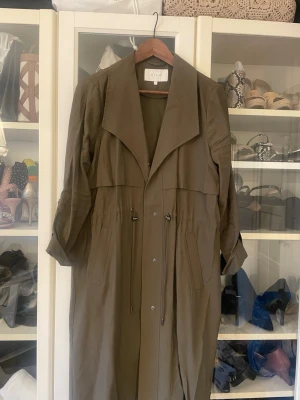 Olivgrön trenchcoat från Vila - Säljer en olivgrön trenchcoat från Vila med stora slag, dragsko i midjan och knappar framtill. Jackan har långa ärmar och två fickor framtill. Perfekt för dig som gillar en clean och avslappnad look.