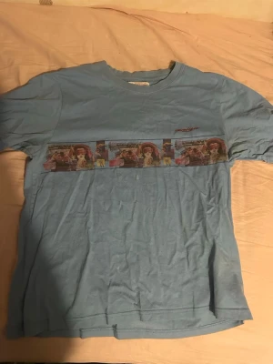 Blå t-shirt med tryck och motiv - Säljer en blå t-shirt med coolt tryck av film- eller seriemotiv som går runt hela tröjan. T-shirten har rund hals och korta ärmar. Perfekt för dig som gillar unika och färgglada plagg med lite retrovibe.