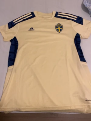 Ingen sko på bilden - Bilden visar ingen sko, utan en gul t-shirt från Adidas med svenska landslagets emblem och blå detaljer på axlar och sidor.