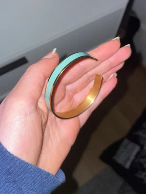 Turkos och guldfärgat armband - Snyggt armband i metall med turkos färg och guldfärgade kanter. Modern och stilren design med en rak, öppen form som gör den enkel att ta på och av. Perfekt accessoar för att lyfta din outfit med en pop av färg. Knappt använd 