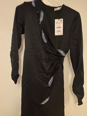 Svart långklänning med blå detaljer från Zara - Säljer en svart långklänning från Zara med snygga blå och silverfärgade broderade detaljer i form av blad. Klänningen har lång ärm, rund hals och en lätt draperad midja. Perfekt för dig som vill ha något unikt och stilrent.