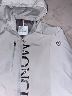 Grå Moncler vindjacka med huva - Snygg grå vindjacka från Moncler med stor logga längs dragkedjan och emblem på ärmen. Storlek L. Pris kan diskuteras vid snabb och smidig affär.