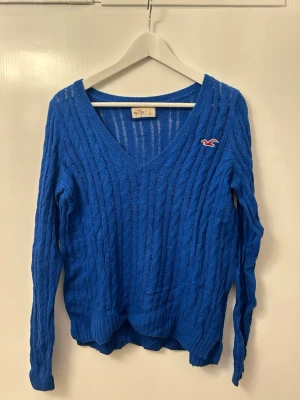 Blå kabelstickad tröja Hollister - Blå kabelstickad tröja från Hollister med v-ringning och lång ärm. Tröjan har ett broderat Hollister-logo på bröstet och är gjord i ett mjukt stickat material. Perfekt för lager-på-lager och chill dagar.
