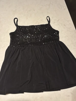 Svart paljettopp från Gina Tricot - Säljer en svart topp från Gina Tricot med smala axelband och paljetter över bysten. Toppen har en glansig, luftig nederdel och är perfekt för dig som vill ha något extra till din outfit. Materialet känns mjukt och toppen har en croppad passform. Använd ungefär 1-3 gånger!💗