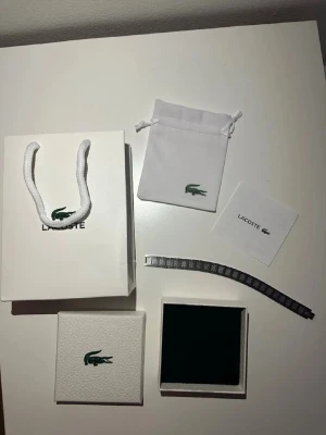 Lacoste silverfärgad armband - Säljer en stilren armbandslänk från Lacoste i silverfärgat metall med graverad Lacoste-logga på spännet. Länken har ett mönstrat rutigt inslag på varje länkdel och kommer med originalförpackning, tygpåse och äkthetsintyg.