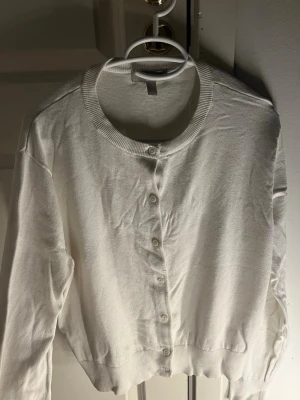 Vit kofta med knappar  - En enkel och stilren vit kofta med rund halsringning och knappar framtill. Koftan har långa ärmar och ribbade muddar vid hals och nederkant. Perfekt att slänga över en topp för en clean look.