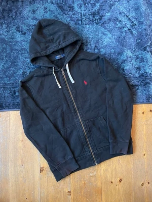 Svart Ralph Lauren Hoodie - Hej, säljer en svart Ralp Lauren zip up Hoodie med en röd pony. Se skick enligt bilder, den har tappat lite färg runt botten av ärmarna, men annars är den riktigt fin. Den kommer i storlek L. Hör av er vid frågor!