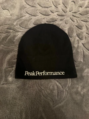 Svart mössa från Peak Performance - En stilren svart mössa från Peak Performance med vit broderad logga framtill. Mössan har en enkel och klassisk form som passar perfekt för kyliga dagar. Materialet känns mjukt och bekvämt mot huvudet.