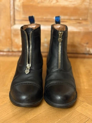 Svarta ridstövletter från Ariat - Snygga svarta stövletter från Ariat i skinn med dragkedja framtill och rund tå. De har en låg klack och robust platt sula, samt blå detaljer på flärpen. Perfekta för ridning och har en stilren look med diskreta sömmar. Kan även användas som vanliga boots. 