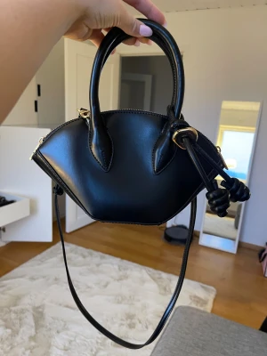 Svart handväska med knutdetaljer - Snygg svart handväska i skinn med unik form och coola knutdetaljer på handtaget. Väskan har guldfärgade metalldetaljer, dragkedja upptill och en axelrem som gör den enkel att bära. Insidan har ett fack med dragkedja för småsaker.💕💕har använt den en gång så den är i nyskick. Man får med dustbag🤩🤩