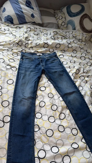 Replay Hyperflex jeans blå 32/32 - Säljer ett par Replay Hyperflex jeans i klassisk blå färg med snygga slitningar och kontrastsömmar. Modellen har fem fickor, normal passform och raka ben. Materialet är stretchigt jeans för extra komfort. Perfekt för dig som gillar stilrena och bekväma jeans.