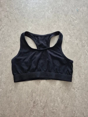 Sport-behå - Svart sportig sport-behå från KappAhl med bred resår nedtill där det står 'BODY ZONE'. Modellen har brottarrygg och är tillverkad i mjuk bomull, perfekt för träning eller chill. Enkel och stilren design som funkar till allt.