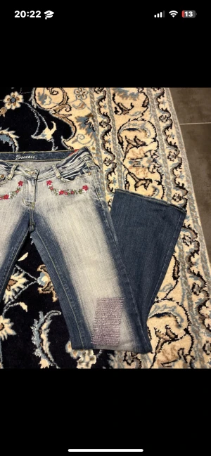 Jeans - Snygga blå jeans med ljus tvätt från Success, dekorerade med broderade blommor i rött, vitt och grönt vid fickorna. Low waist. Jeansen har raka ben och patchade detaljer på låren. Finns enstaka små fläckar (ej märkbart) men kan försvinna med tvätt. Passar XS-S. L32