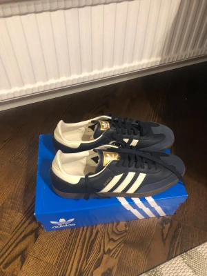 Adidas Samba sneakers marinblå/vit - Adidas Samba sneakers i marinblått och vitt med klassiska tre ränder på sidan, guldiga detaljer på tungan och texten 'SAMBA' i guld. Ovandel i skinn och mocka, platt brun gummisula och snörning. Aldrig använda. De är i storlek US 13, och är i EU 48. Däremot mycket små i storleken så passar nog 46-47.   Helt nya, med låda. Skriv vid frågor