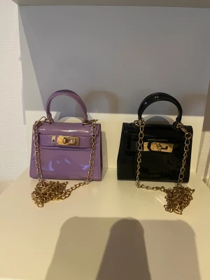 Lila och svart minihandväska med kedja - Två små handväskor i lila och svart med glansig finish, guldfärgade kedjor och låsdetaljer. Perfekt accessoar för att lyfta din outfit med en trendig touch. Väskorna har ett stilrent och kompakt format med handtag upptill. Den säljs båda för 100kr och en för 50kr.