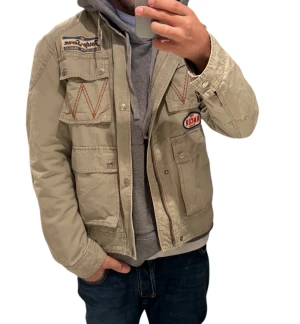 Beige militär jacka  - Säljer en beige parkajacka från Rager med coola patchar och broderade detaljer på bröstet och ärmen. Jackan har flera fickor med knappar och dragkedja framtill. Perfekt för dig som gillar och vill sticka ut lite extra.